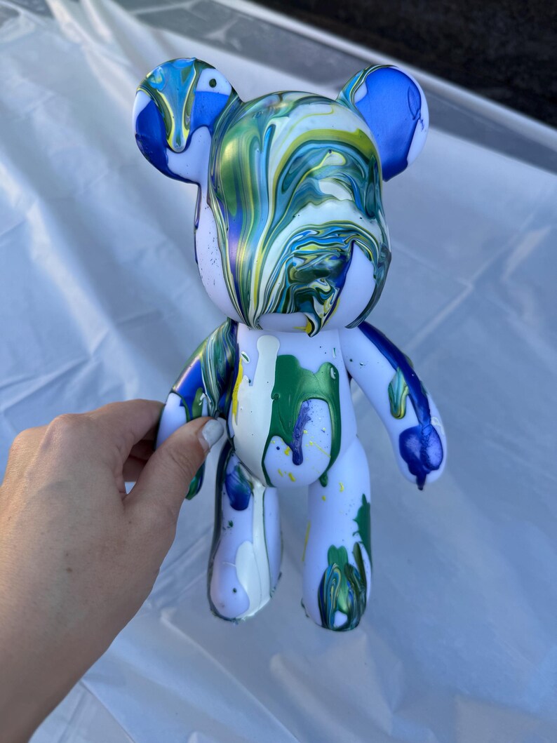 May include: A bear-shaped figurine with a white base color, featuring a fluid paint pour design in shades of blue, green, and yellow. The paint drips down from the head and body, creating a marbled, glossy effect across the surface.