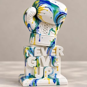 May include: A white boxing glove sculpture featuring blue, green, and yellow paint drip patterns. The glove rests on a rectangular base with the raised white text NEVER GIVE UP, also accented with blue, green, and yellow paint drips.