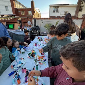 May include: A group of children gathered around a long white table outdoors, painting small white bear-shaped figurines with various colorful paints from small squeeze bottles. Each child has a paper plate holding their figurine and paint.
