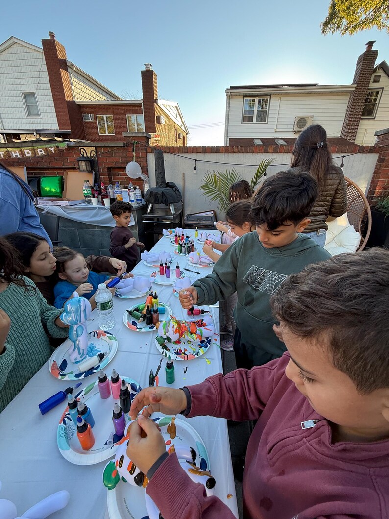 May include: A group of children gathered around a long white table outdoors, painting small white bear-shaped figurines with various colorful paints from small squeeze bottles. Each child has a paper plate holding their figurine and paint.