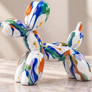 May include: A glossy white balloon dog sculpture featuring a vibrant multi-colored paint drip pattern in shades of blue, green, orange, and purple. The figurine is shaped like a twisted balloon animal with a rounded head, body, and four legs.