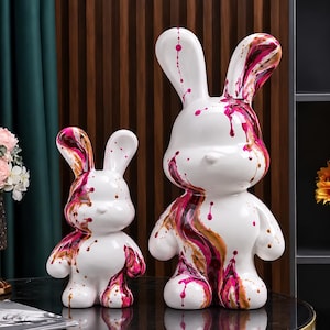 May include: Two white rabbit figurines of different sizes, each featuring a glossy finish with abstract paint drips in shades of magenta, dark red, and metallic gold. The sculptures have smooth, rounded forms and long ears.