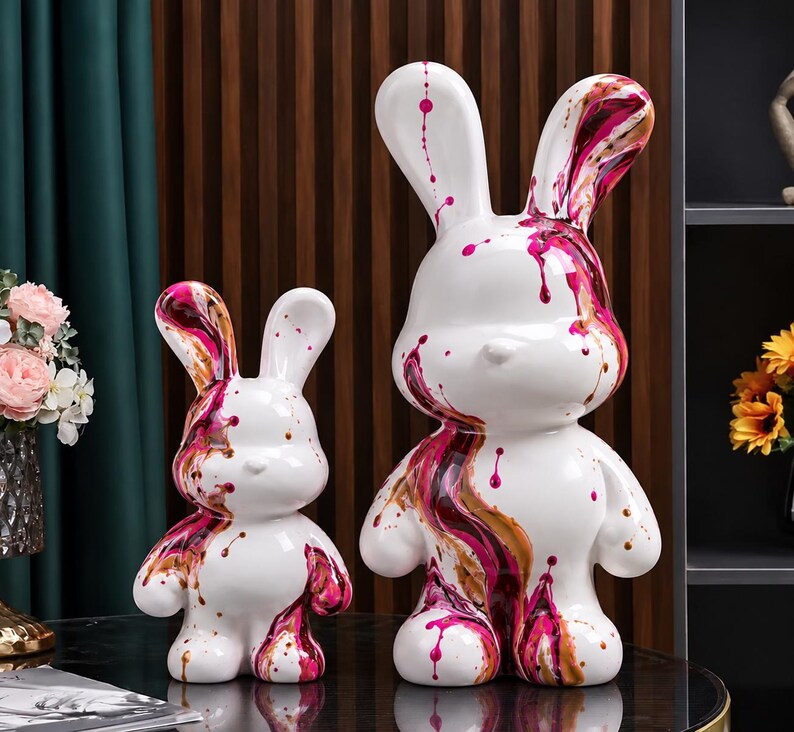 May include: Two white rabbit figurines of different sizes, each featuring a glossy finish with abstract paint drips in shades of magenta, dark red, and metallic gold. The sculptures have smooth, rounded forms and long ears.