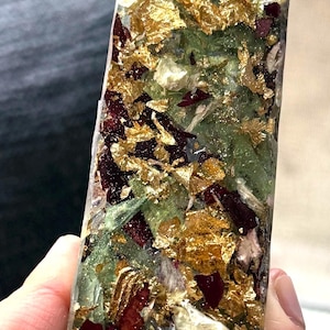 May include: A rectangular resin mezuzah case featuring a clear finish embedded with dried dark red flower petals, green leaves, and gold leaf flakes. An orange Hebrew letter Shin is prominently attached to the top portion of the front surface.