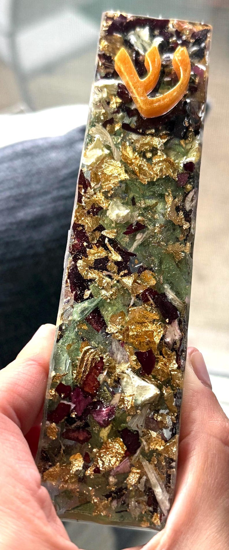 May include: A rectangular resin mezuzah case featuring a clear finish embedded with dried dark red flower petals, green leaves, and gold leaf flakes. An orange Hebrew letter Shin is prominently attached to the top portion of the front surface.