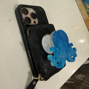 May include: Black phone case with a quilted texture and a zipper pocket. A blue and turquoise octopus-shaped phone grip is attached. The phone case has a gold-colored zipper and a black wrist strap.