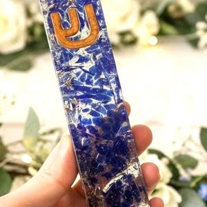 May include: A rectangular mezuzah case made of clear resin embedded with deep blue lapis lazuli stone fragments and metallic gold leaf flakes. The top section features a raised, light brown wooden Hebrew letter Shin centered on the surface.