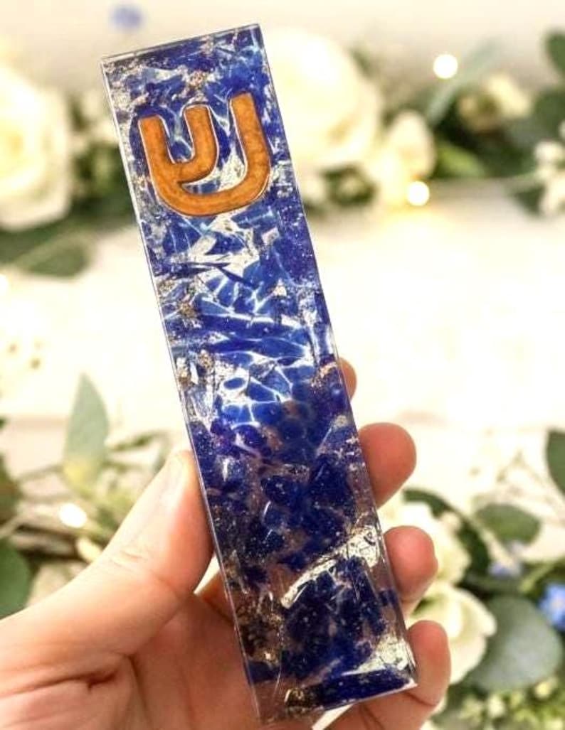 May include: A rectangular mezuzah case made of clear resin embedded with deep blue lapis lazuli stone fragments and metallic gold leaf flakes. The top section features a raised, light brown wooden Hebrew letter Shin centered on the surface.