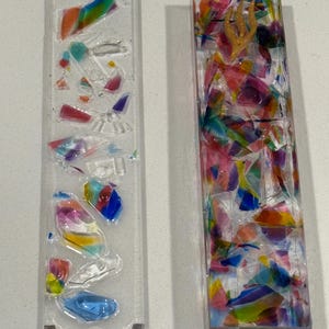 May include: Two rectangular mezuzah cases made of clear resin. The left case is flat with embedded colorful glass shards. The right case is a three-dimensional block filled with multicolored glass fragments and a gold-colored Hebrew letter shin.