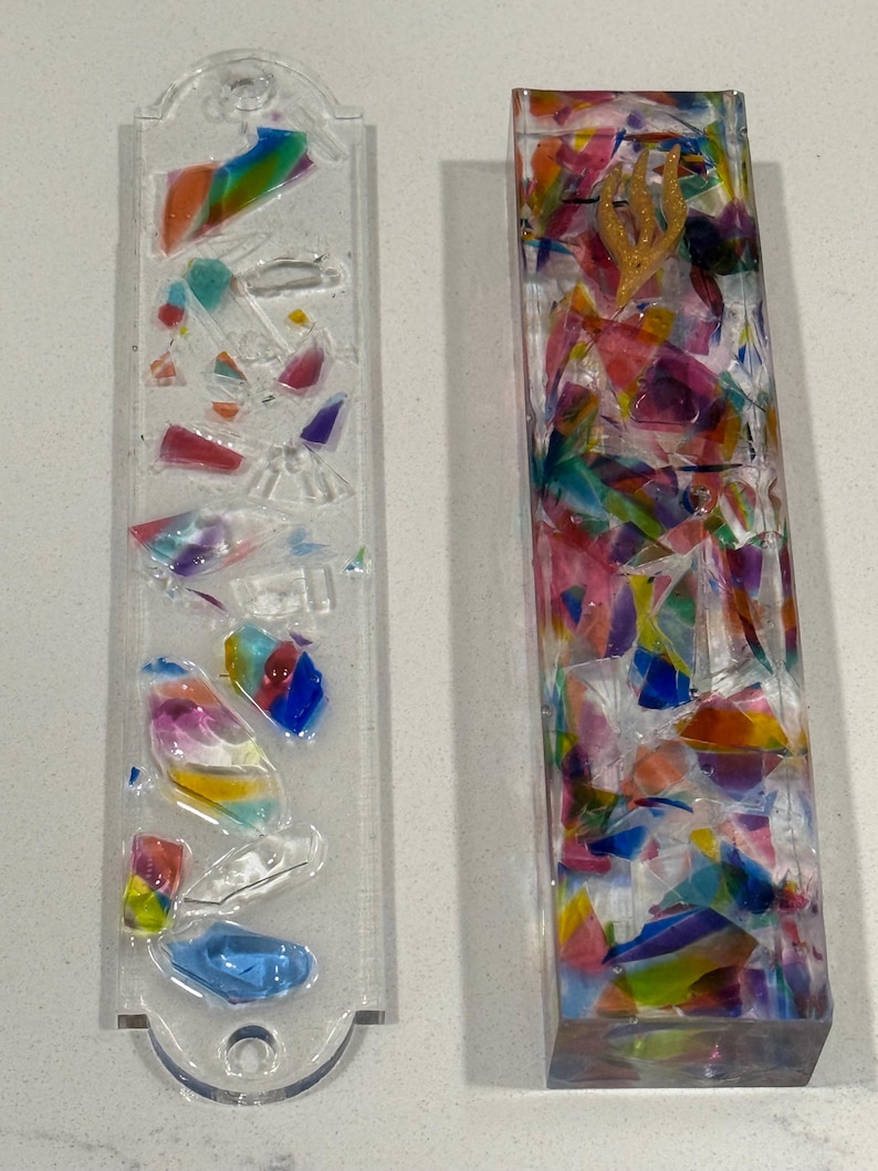 May include: Two rectangular mezuzah cases made of clear resin. The left case is flat with embedded colorful glass shards. The right case is a three-dimensional block filled with multicolored glass fragments and a gold-colored Hebrew letter shin.