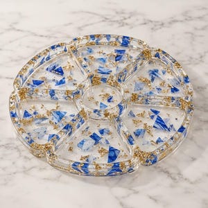 May include: A circular resin serving tray featuring six wedge-shaped compartments and a central circular section. The clear resin is embedded with small, angular pieces of blue and white marbled material and shimmering gold leaf flakes throughout.