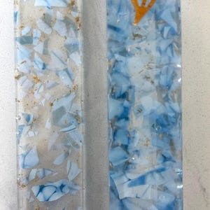 May include: Two rectangular resin mezuzah cases featuring a clear base embedded with light blue and white shards and gold leaf flakes. The left case has rounded ends with holes, while the right case features a gold-colored shin symbol at the top.