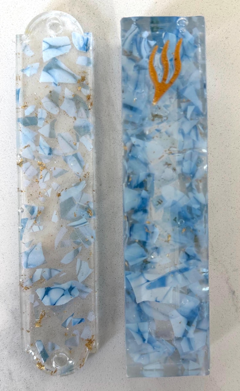 May include: Two rectangular resin mezuzah cases featuring a clear base embedded with light blue and white shards and gold leaf flakes. The left case has rounded ends with holes, while the right case features a gold-colored shin symbol at the top.