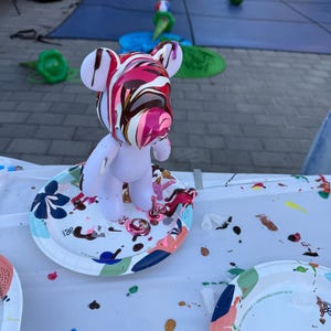 May include: A white bear-shaped figurine standing on a patterned paper plate. The head of the figurine is covered in a thick, glossy pour of swirling pink, white, and dark brown paint that drips down the sides of the face and ears.