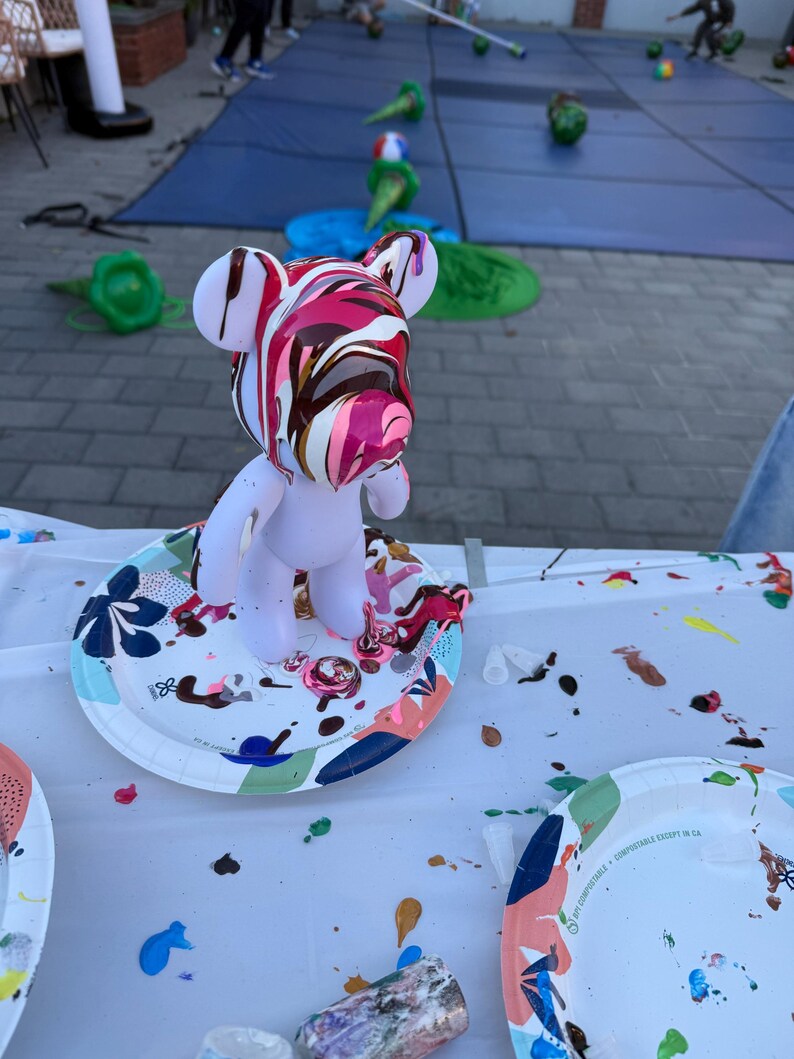 May include: A white bear-shaped figurine standing on a patterned paper plate. The head of the figurine is covered in a thick, glossy pour of swirling pink, white, and dark brown paint that drips down the sides of the face and ears.
