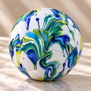 May include: A soccer ball featuring a white base with a vibrant, marbled paint design in shades of blue, teal, and yellow. The surface displays fluid, swirling patterns of color that drip across the panels, creating a dynamic and colorful appearance.