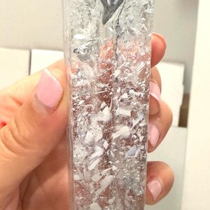 May include: A rectangular, clear resin mezuzah case featuring embedded silver-toned metallic flakes and white fragments throughout the body. A raised, silver-colored Hebrew letter Shin is positioned near the top of the front face of the transparent object.