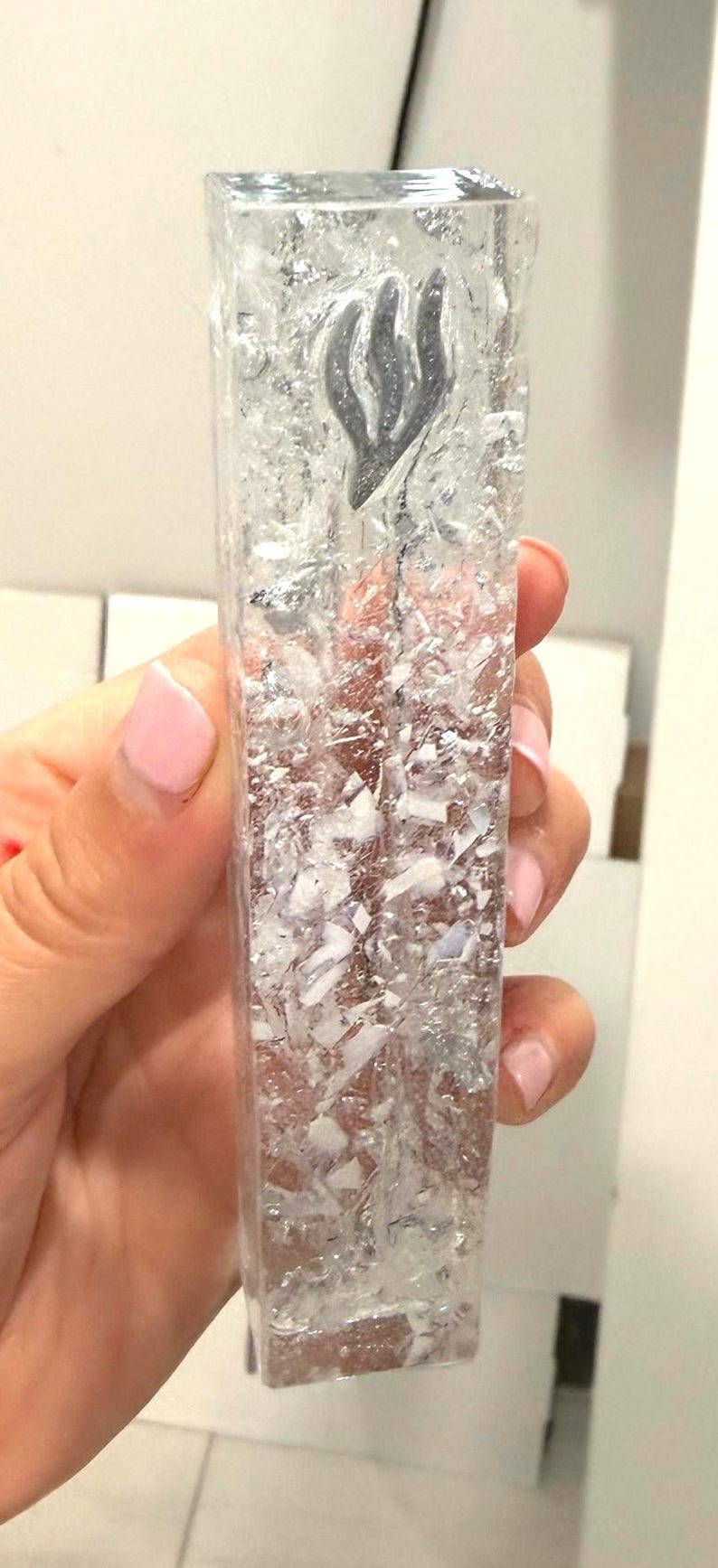 May include: A rectangular, clear resin mezuzah case featuring embedded silver-toned metallic flakes and white fragments throughout the body. A raised, silver-colored Hebrew letter Shin is positioned near the top of the front face of the transparent object.