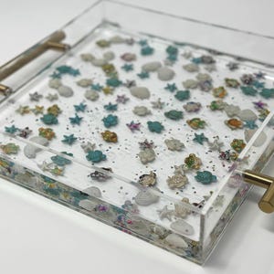 May include: Clear acrylic tray with gold handles, filled with miniature sea creatures and glitter. The tray features a variety of colorful turtles, starfish, and shells, creating a coastal theme. The tray is square-shaped.