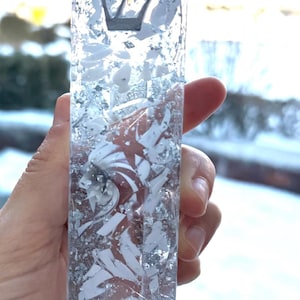 May include: A rectangular mezuzah case made of clear resin embedded with silver metallic flakes and white shards. The Hebrew letter Shin is prominently displayed in black near the top of the vertical, transparent, and textured decorative object.