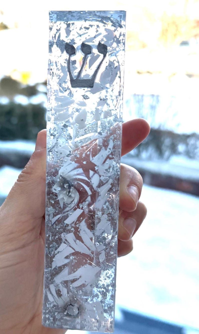 May include: A rectangular mezuzah case made of clear resin embedded with silver metallic flakes and white shards. The Hebrew letter Shin is prominently displayed in black near the top of the vertical, transparent, and textured decorative object.