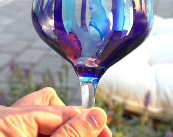 May include: A clear glass wine glass featuring vertical abstract drips of blue, light blue, and metallic gold paint. The stem and circular base are accented with a deep blue tint, creating a vibrant contrast against the transparent glass bowl.