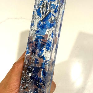 May include: A rectangular mezuzah case made of clear resin embedded with deep blue glass shards and metallic silver leaf flakes. A silver-toned Hebrew letter Shin is affixed to the upper portion of the front face of the elongated, vertical object.