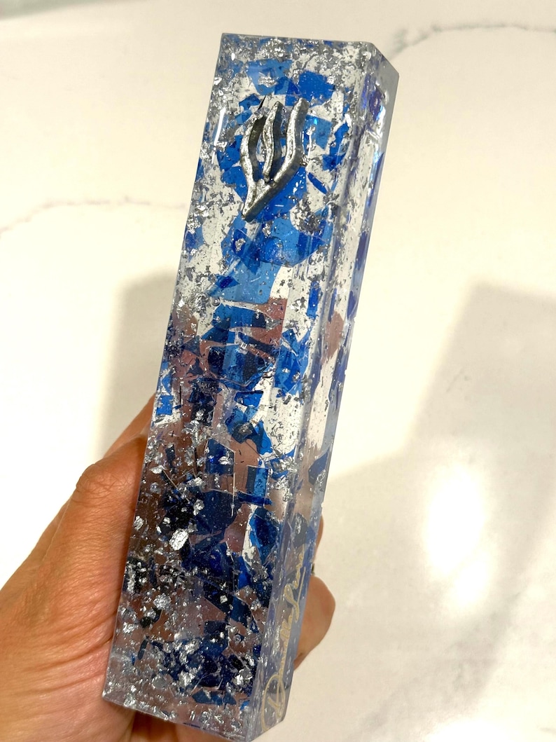 May include: A rectangular mezuzah case made of clear resin embedded with deep blue glass shards and metallic silver leaf flakes. A silver-toned Hebrew letter Shin is affixed to the upper portion of the front face of the elongated, vertical object.