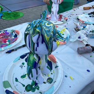 May include: A light purple bear-shaped figurine covered in dripping, swirled paint in shades of blue, green, and yellow. The figurine stands on a white paper plate with colorful abstract patterns, surrounded by paint supplies and another figurine.