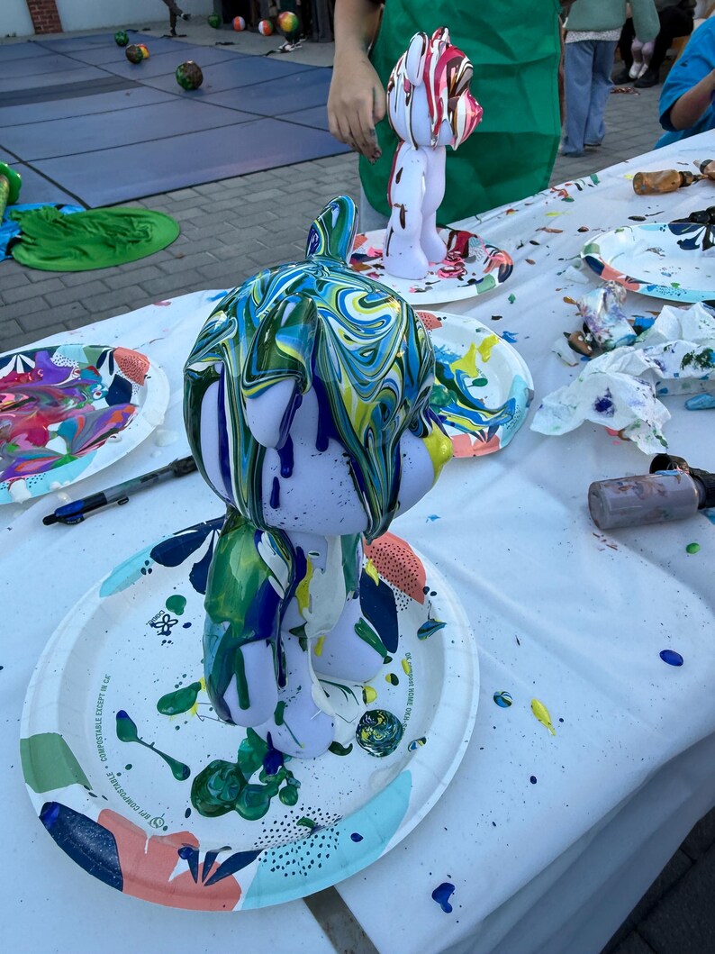 May include: A light purple bear-shaped figurine covered in dripping, swirled paint in shades of blue, green, and yellow. The figurine stands on a white paper plate with colorful abstract patterns, surrounded by paint supplies and another figurine.