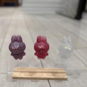 May include: Three glittery bunny figurines in purple, red, and white, each with a sparkling finish. The bunnies are displayed on a small wooden stand, showcasing their unique colors and textures. These decorative items are ideal for Easter or spring decor.