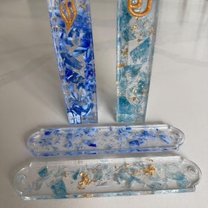 May include: Two rectangular resin mezuzah cases in shades of blue and white with gold leaf accents and a gold Hebrew letter Shin on each. Two matching flat rectangular resin plates with blue and light blue fragments and gold leaf flakes.
