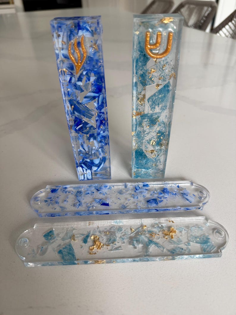 May include: Two rectangular resin mezuzah cases in shades of blue and white with gold leaf accents and a gold Hebrew letter Shin on each. Two matching flat rectangular resin plates with blue and light blue fragments and gold leaf flakes.