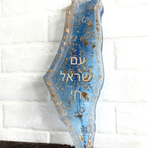 May include: A blue resin art piece in the shape of a map, with gold flakes and Star of David accents. The piece has Hebrew text. The background is a white brick wall.