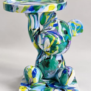 May include: A decorative bear-shaped side table featuring a glossy finish with a vibrant, multi-colored paint drip pattern in shades of blue, green, yellow, purple, and white. The bear sits on a square base and holds a circular tray overhead.
