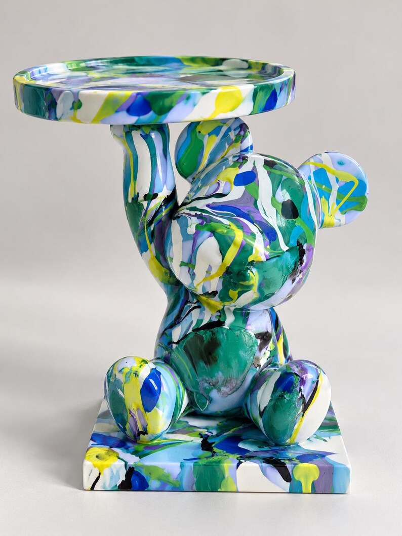 May include: A decorative bear-shaped side table featuring a glossy finish with a vibrant, multi-colored paint drip pattern in shades of blue, green, yellow, purple, and white. The bear sits on a square base and holds a circular tray overhead.