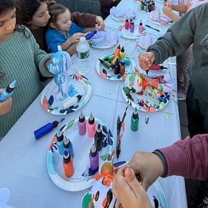 May include: A group of individuals participating in a craft activity, painting white bear-shaped figurines with colorful acrylic paint from squeeze bottles. The figurines are placed on patterned paper plates, with various paint colors scattered across the table.