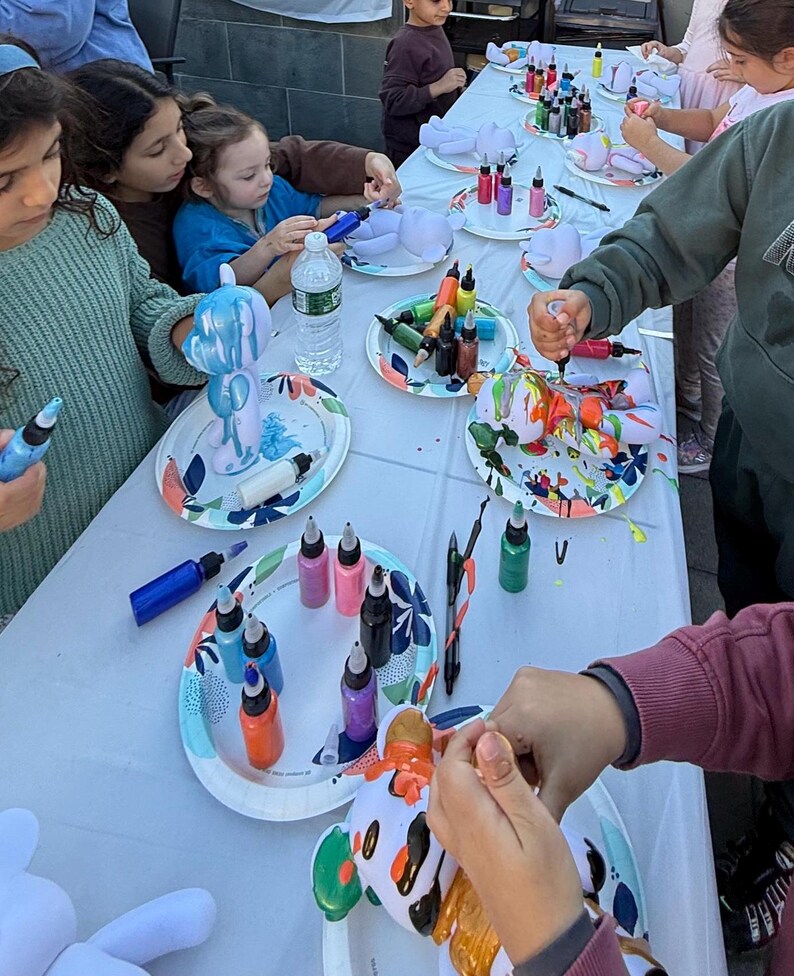 May include: A group of individuals participating in a craft activity, painting white bear-shaped figurines with colorful acrylic paint from squeeze bottles. The figurines are placed on patterned paper plates, with various paint colors scattered across the table.