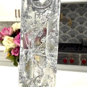 May include: A rectangular clear resin mezuzah case featuring embedded silver leaf flakes and a raised silver Hebrew letter Shin at the top. The transparent material displays a textured, reflective appearance with visible air bubbles throughout the structure.