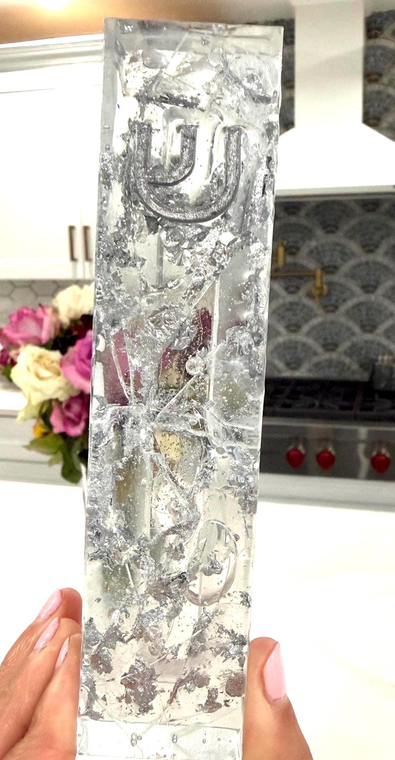 May include: A rectangular clear resin mezuzah case featuring embedded silver leaf flakes and a raised silver Hebrew letter Shin at the top. The transparent material displays a textured, reflective appearance with visible air bubbles throughout the structure.