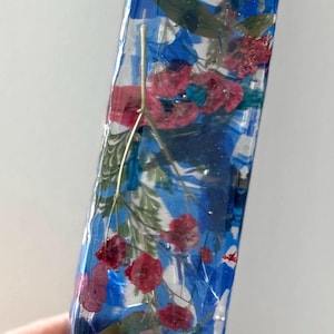 May include: A rectangular mezuzah case made of clear resin, featuring embedded dried red and green flowers and blue pigment swirls. A gold-colored Hebrew letter Shin is attached to the upper portion of the front surface of the transparent casing.
