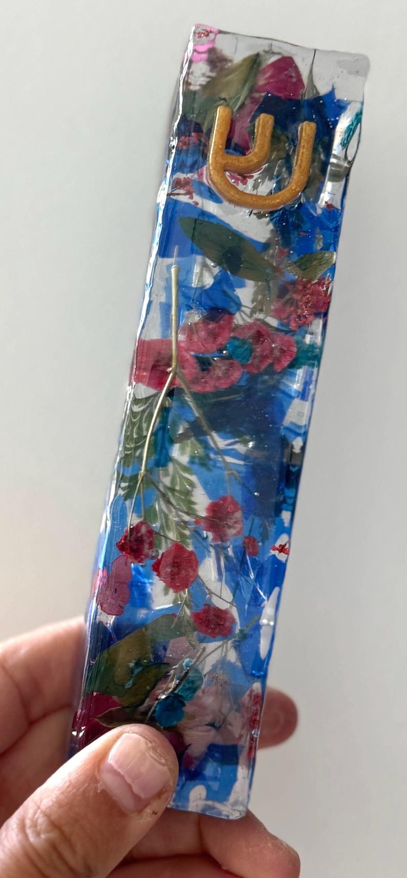 May include: A rectangular mezuzah case made of clear resin, featuring embedded dried red and green flowers and blue pigment swirls. A gold-colored Hebrew letter Shin is attached to the upper portion of the front surface of the transparent casing.