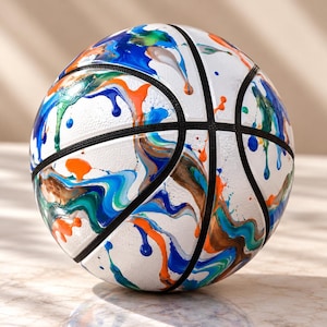 May include: A white basketball featuring a textured surface with abstract paint drips in shades of blue, green, orange, and brown. The ball includes standard black rubber seams that divide the surface into panels with a colorful pattern.