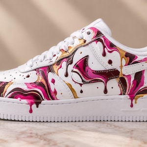 May include: A white sneaker featuring a vibrant paint drip design in shades of hot pink, metallic gold, and deep chocolate brown. The shoe has white laces, a white midsole, and a white outsole, with the colorful liquid pattern covering the side panels.