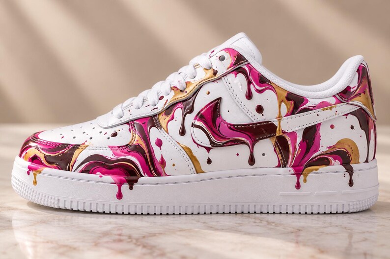 May include: A white sneaker featuring a vibrant paint drip design in shades of hot pink, metallic gold, and deep chocolate brown. The shoe has white laces, a white midsole, and a white outsole, with the colorful liquid pattern covering the side panels.