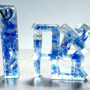 May include: A vertical rectangular resin mezuzah case featuring a black Hebrew letter Shin and a separate resin sculpture displaying the Hebrew word Chai, both filled with clear resin and embedded with fragments of blue and white glass.