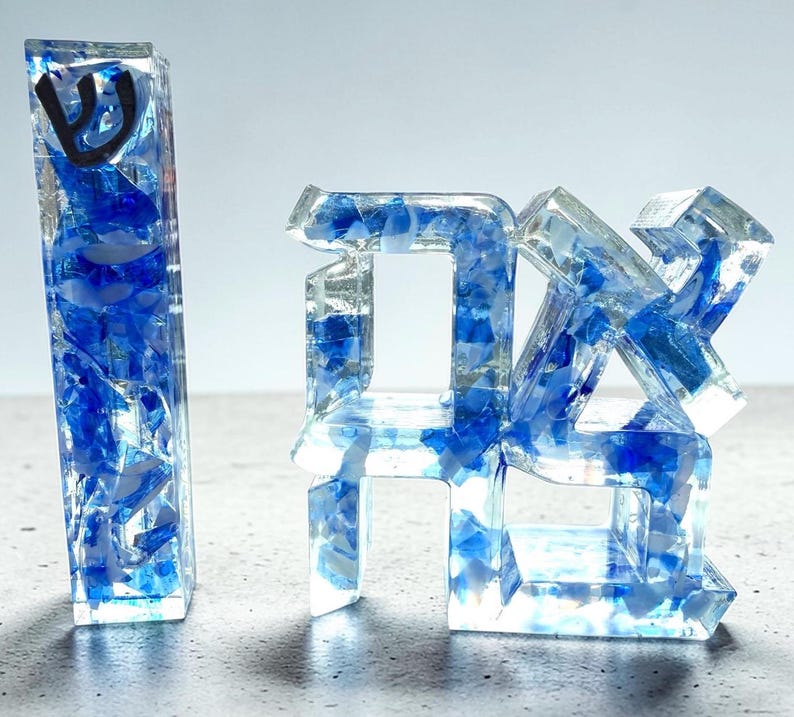 May include: A vertical rectangular resin mezuzah case featuring a black Hebrew letter Shin and a separate resin sculpture displaying the Hebrew word Chai, both filled with clear resin and embedded with fragments of blue and white glass.