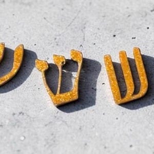 May include: Five gold-colored, glittery, U-shaped decorative pieces arranged in a row. Each piece features three vertical prongs connected at the base, resembling the Hebrew letter Shin, with a textured, sparkling finish and distinct shadows.