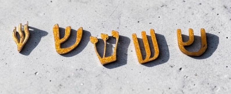 May include: Five gold-colored, glittery, U-shaped decorative pieces arranged in a row. Each piece features three vertical prongs connected at the base, resembling the Hebrew letter Shin, with a textured, sparkling finish and distinct shadows.
