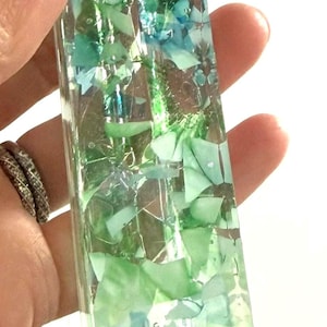 May include: A rectangular mezuzah case made of clear resin embedded with various shades of green and blue glass fragments. The top section features a dark gray Hebrew letter Shin, which is a traditional symbol found on this type of religious object.